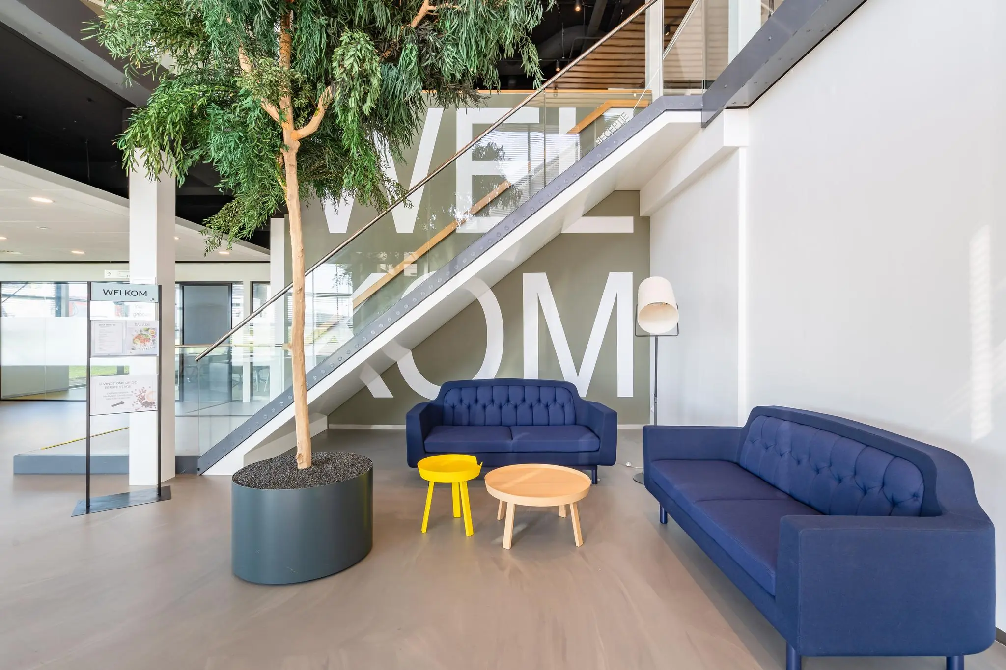 Moderne ontvangstruimte in Rivium Promenade met blauwe banken, een grote plant en de tekst 'WELKOM' op de muur.